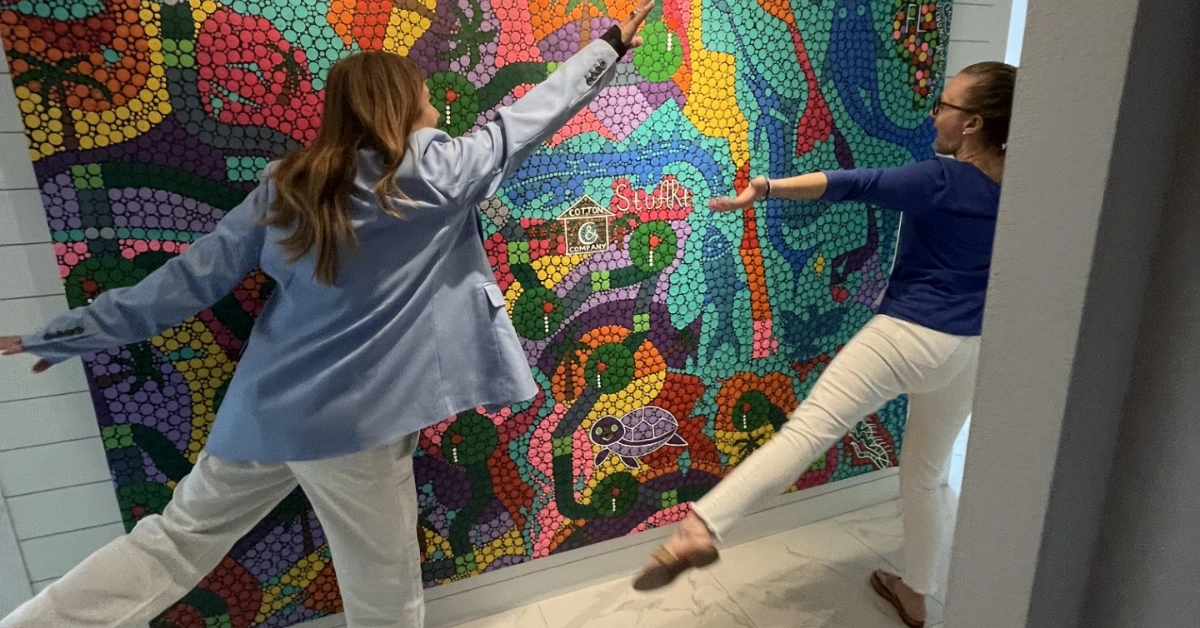 Two women pose in front of colorful mosaic wall artwork indoors