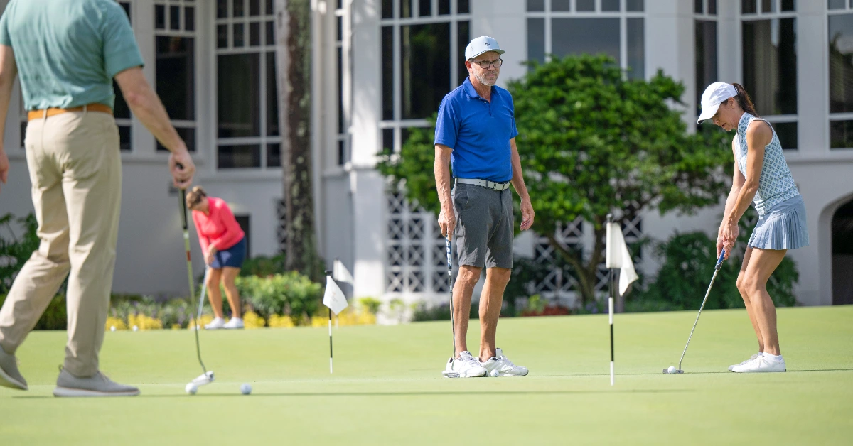 Four golfers practice putting on a green near a building with large windows.