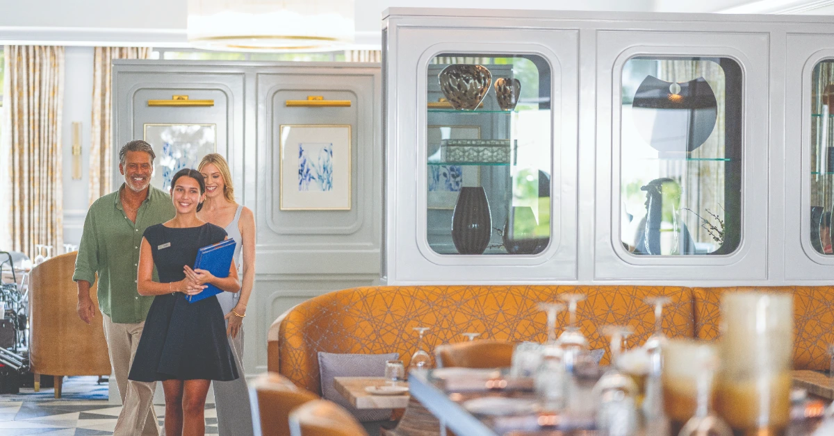 Staff member leads a couple through an elegant dining room with booth seating and display cabinets
