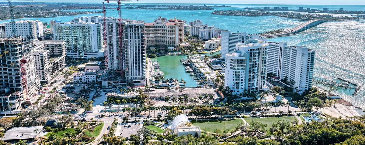 Aerial harbor view with waterfront towers, cranes, marina channels, parkland, and a long bridge over blue water.