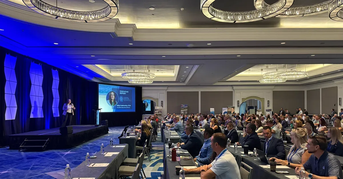 Kelly Grabowsky, CCM, presents the Executive Report to hundreds of attendees at a packed real estate conference in an elegant hotel ballroom.