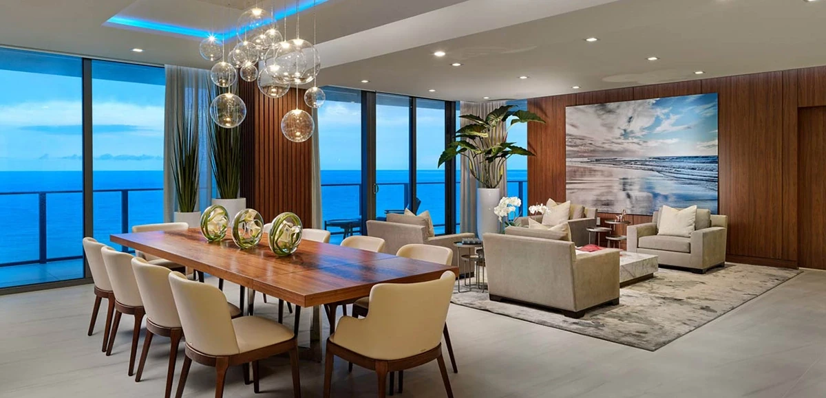 Elegant oceanfront living and dining space featuring a walnut dining table, globe pendant lights, and floor-to-ceiling windows with sweeping sea views.