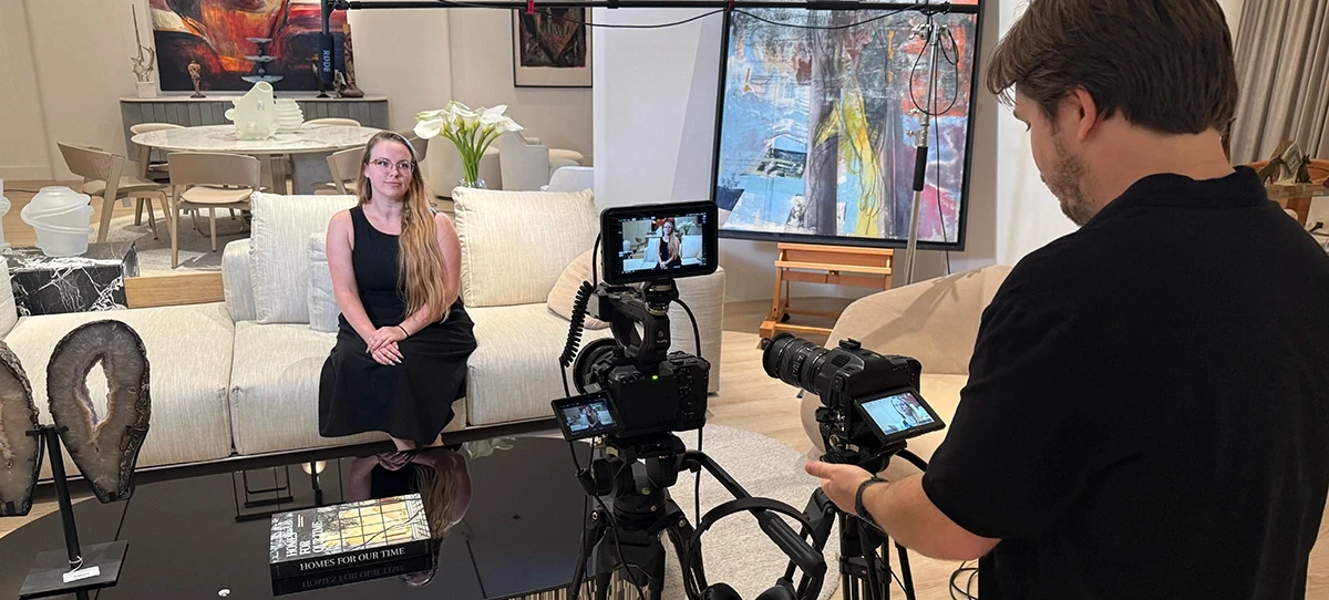 A videographer films an interview with a woman seated on a white sofa in a stylishly furnished modern real estate showroom.